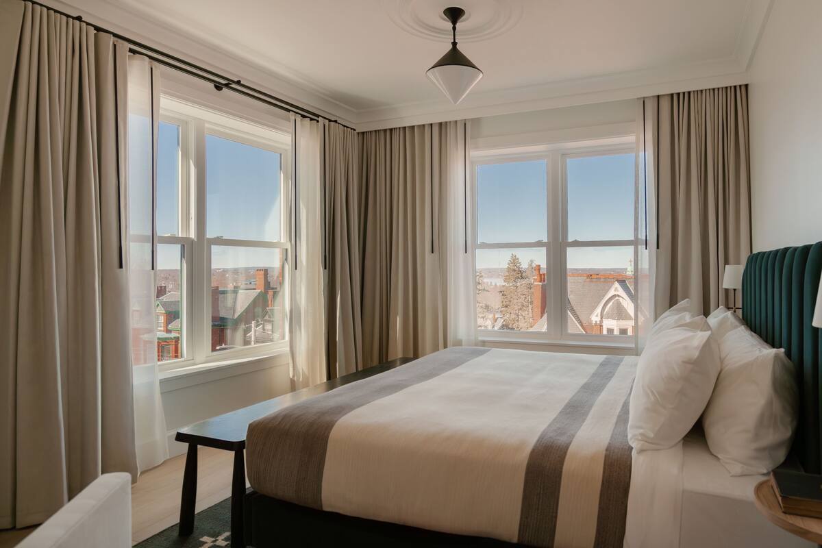 Ein Hotelzimmer mit Bett, Nachttisch, Lampe und Blick auf die Stadt durch große Fenster.