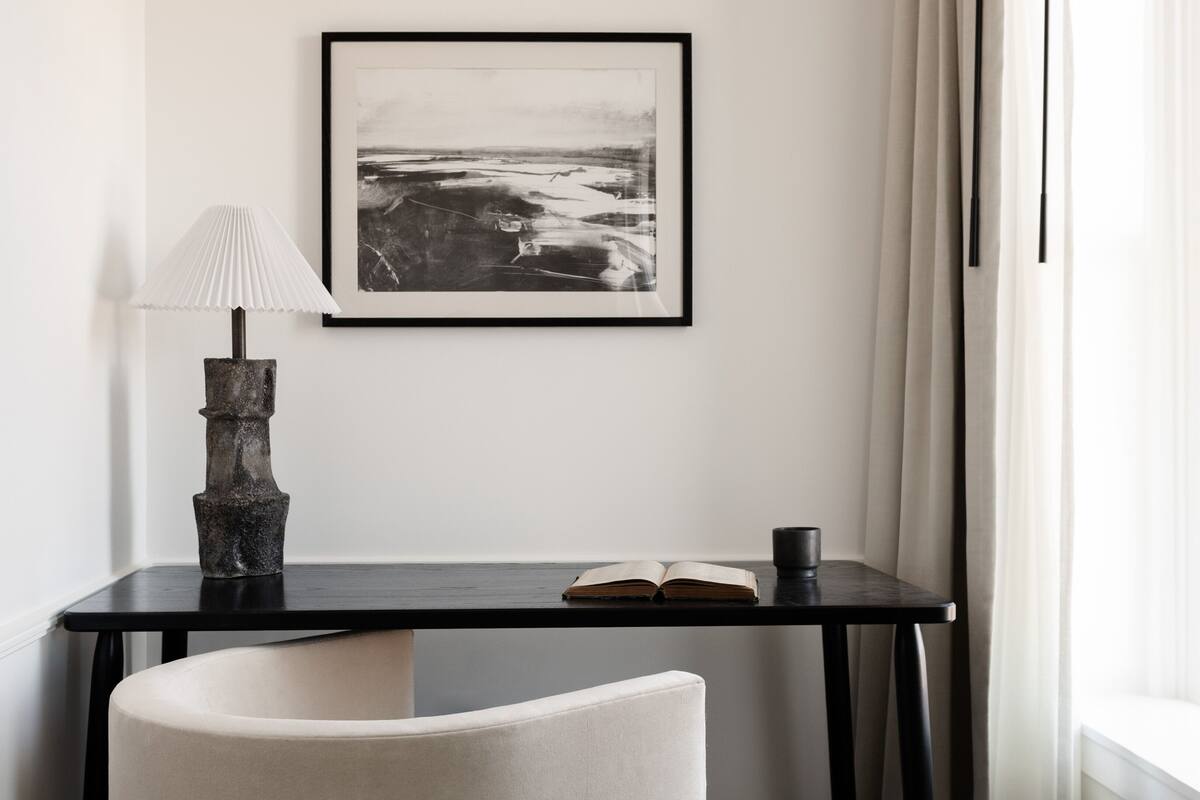 Ein Zimmer mit einem schwarzen Tisch, einem weißen Stuhl, einer Lampe, einem offenen Buch und einem gerahmten Bild an der Wand.