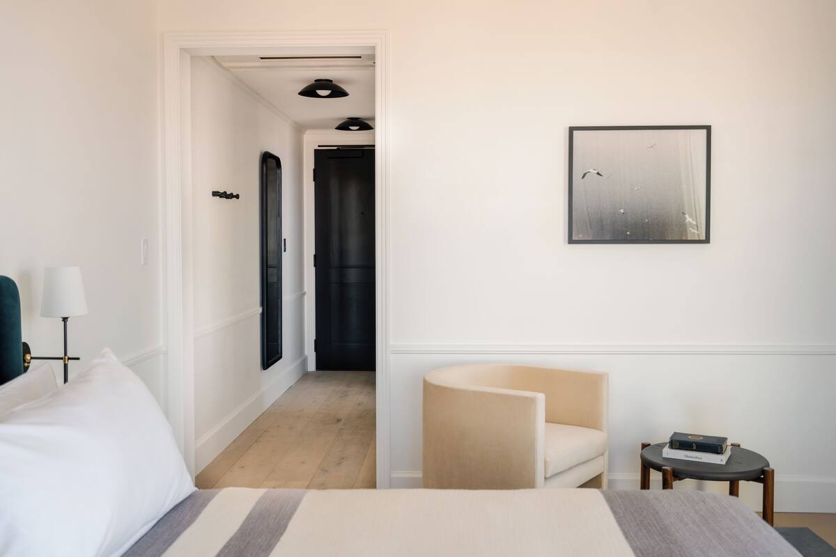 Ein Hotelzimmer mit einem Bett, einem Sessel, einem kleinen Tisch, einer Lampe, einem gerahmten Bild und einer Tür zu einem Flur.