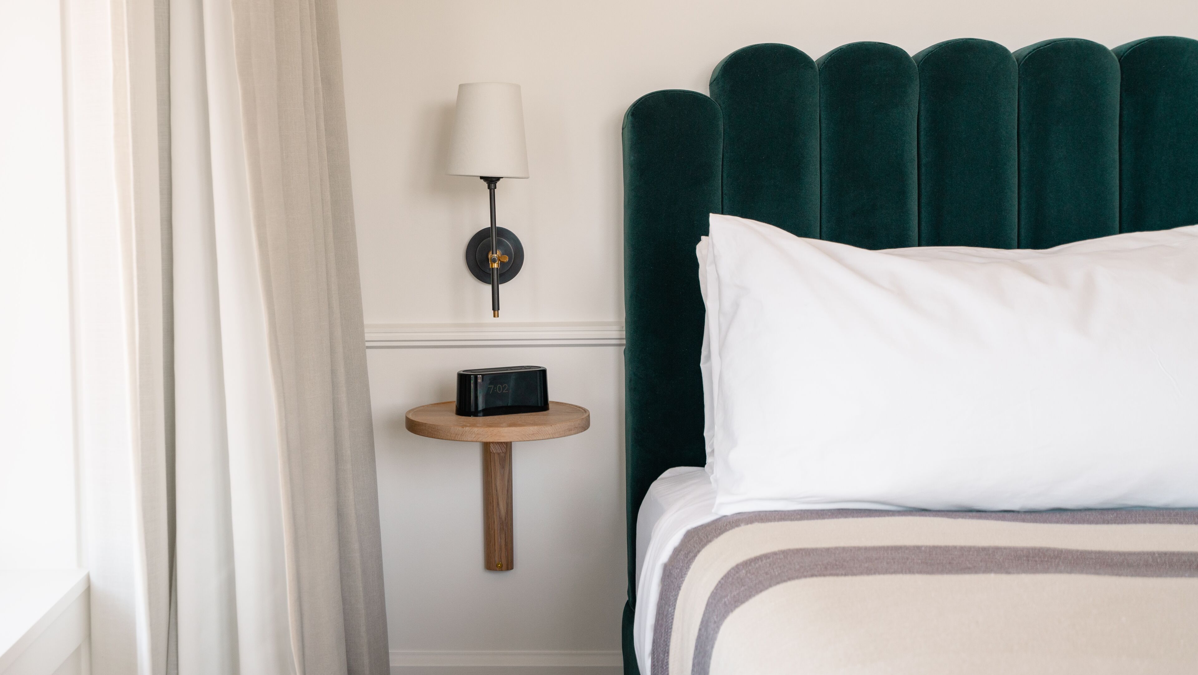 A neatly made bed with a striped pillow and a dark green headboard. A small wooden nightstand with a lamp and a clock is beside the bed.