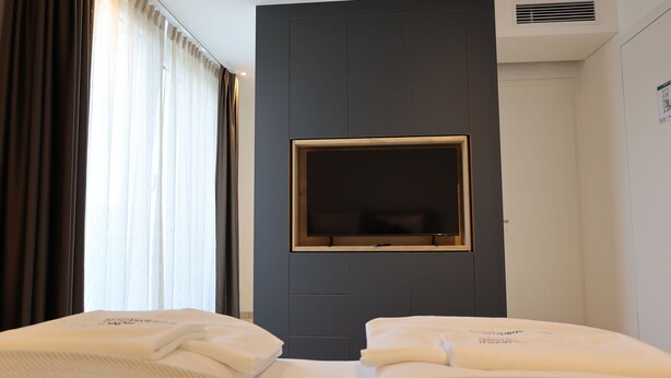 A hotel room with a large TV mounted on a dark wall, two beds with white linens, and a window with curtains. - 1 Doppelbett Zimmer 21 (München)