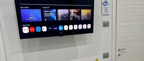 Una habitación moderna con un televisor de montaje que muestra diferentes aplicaciones de streaming, una mesa blanca con platos decorativos, una planta pequeña y una silla blanca.