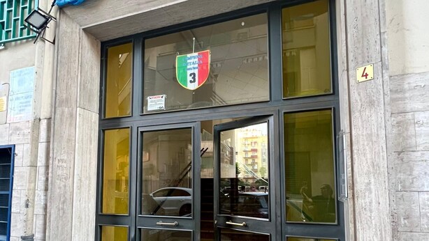Un bâtiment doté d’une grande entrée en verre, d’un escalier visible à l’intérieur et d’une enseigne avec un emblème en forme de blason au-dessus de la porte.