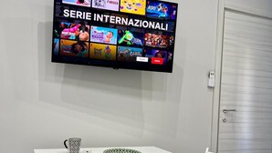 Une pièce avec une table blanche, deux chaises, un téléviseur diffusant différentes émissions et une porte avec un téléphone.