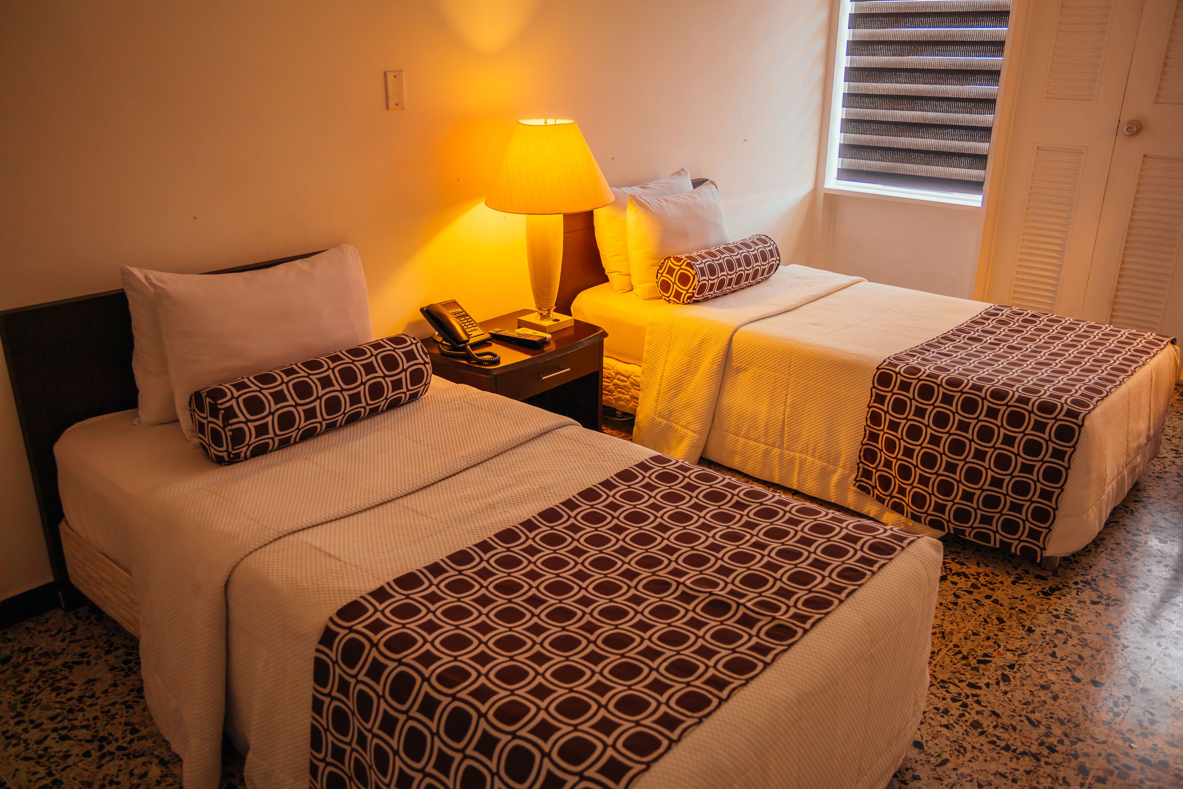Habitación de hotel con dos camas, una mesita de noche con lámpara y una ventana con persianas.