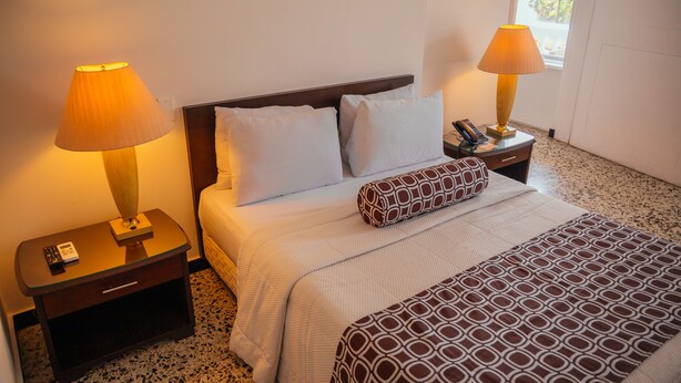 A hotel room with a bed, two bedside lamps, a nightstand with a phone, and a window with a view of palm trees. - Hotel Estación by Destino Pacifico (Buenaventura)