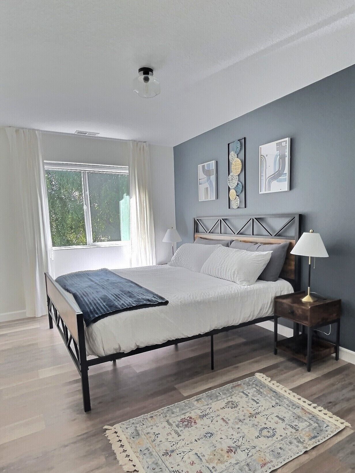 Une chambre à coucher comprenant un lit, une table de chevet, une lampe, une fenêtre avec des rideaux et des œuvres d’art murales.