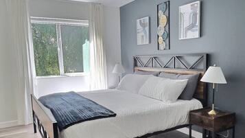 Une chambre à coucher comprenant un lit, une table de chevet, une lampe, une fenêtre avec des rideaux et des œuvres d’art murales.