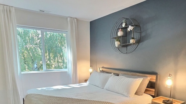 Une chambre moderne avec un grand lit, une table de chevet avec une lampe et une vue sur les arbres par la fenêtre.