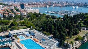 Vista aerea di una zona costiera con un porto turistico, una piscina e un edificio di grandi dimensioni dal design moderno.