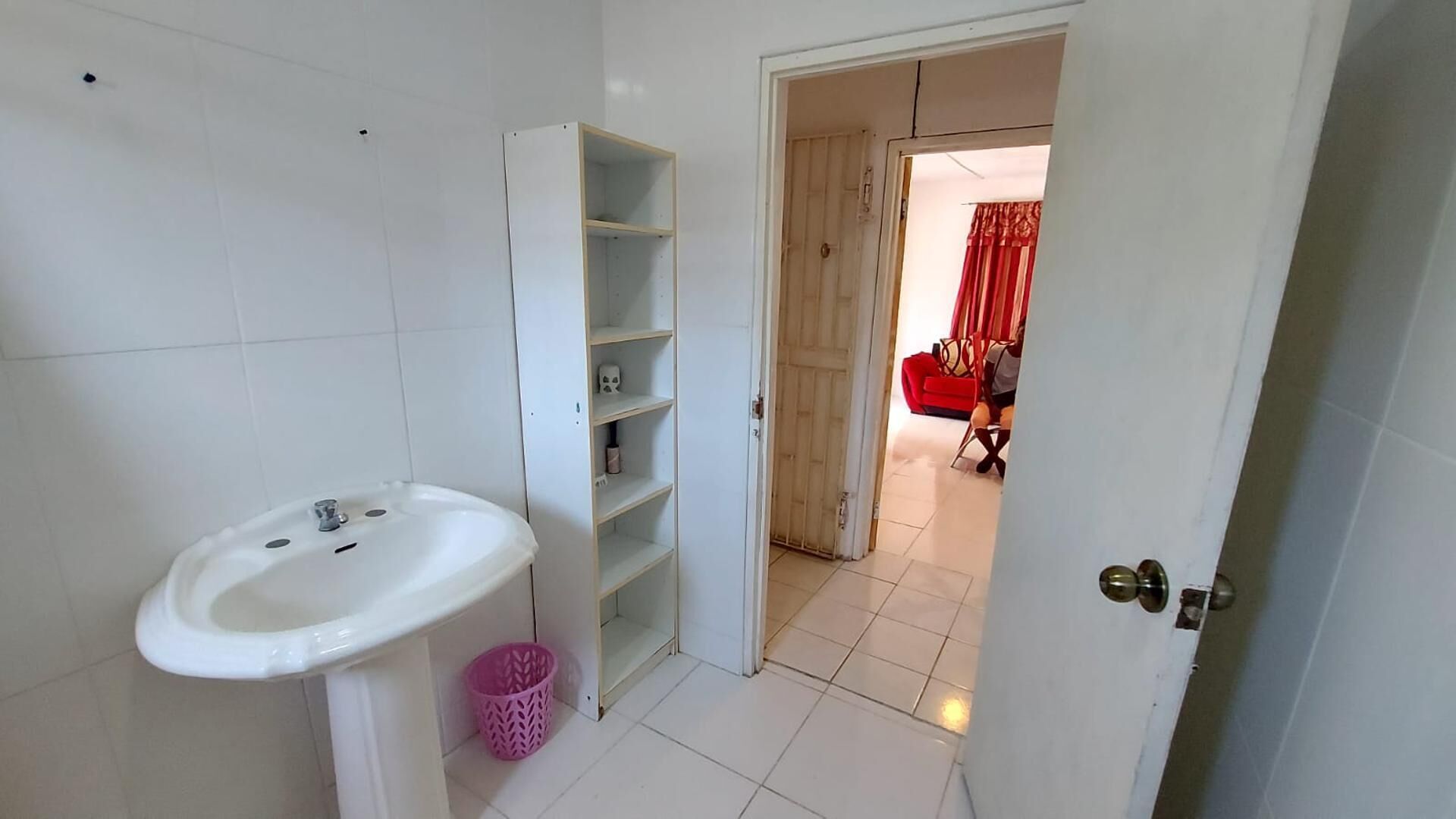 A bathroom with a white sink, a pink basket, and a wooden door leading to a living area with a red sofa.
