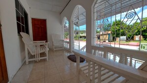 A spacious room with white walls, a tiled floor, and large arched windows with metal grills. There are two white chairs and a wooden bench. - House - Sleeps 6 - Patio - Parking (Port Morant)