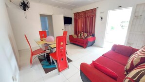 A living room with a red sofa, a dining area with a glass table and red chairs, and a TV mounted on the wall. - House - Sleeps 6 - Patio - Parking (Port Morant)