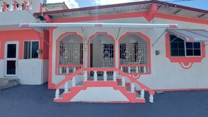 A two-story house with a red and white exterior, a front porch with a white balustrade, and three arched windows with decorative metal grilles. - House - Sleeps 6 - Patio - Parking (Port Morant)