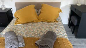 Una cama bien hecha con un edredón estampado floral, dos almohadas de color mostaza y dos toallas dobladas sobre un banco de madera.