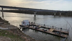 Een steiger aan de rivier met een klein wit gebouw, mensen die wandelen en een brug op de achtergrond.