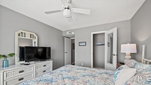 A bedroom with a bed, dresser, TV, ceiling fan, and a door leading to another room.