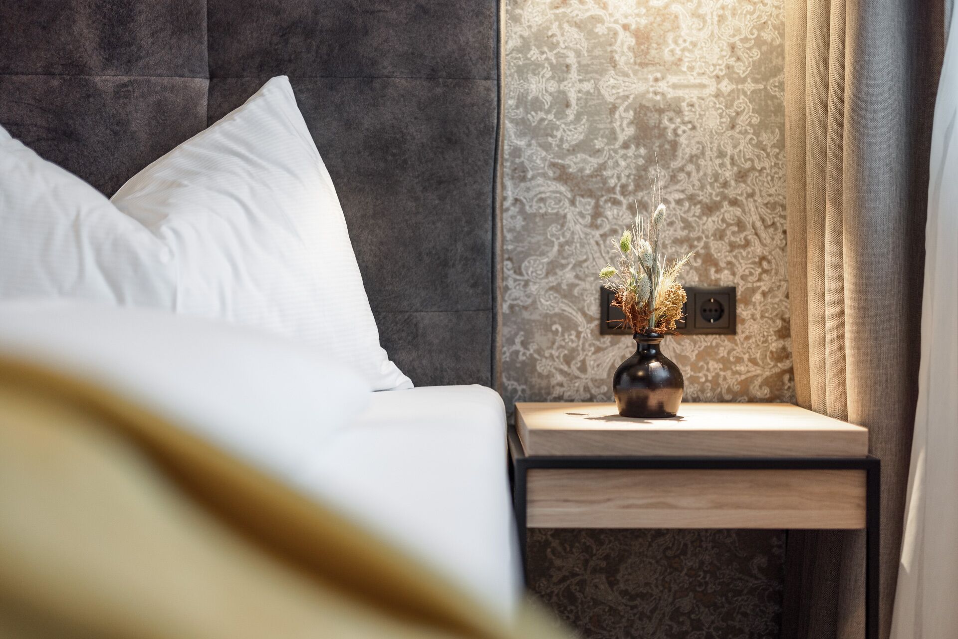 A neatly made bed with white pillows, a bedside table with a vase of dried flowers, and a patterned wall behind.