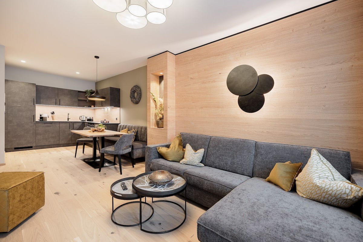 Ein modernes Wohnzimmer mit grauem Sofa, rundem Couchtisch und einem Essbereich, im Hintergrund eine Küche.