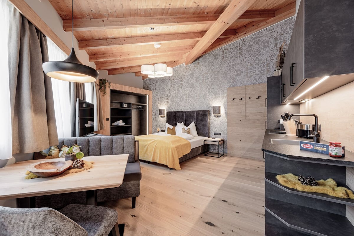 Ein modernes Hotelzimmer mit Holzboden, einem Bett mit dunklem Kopfteil, einem Essbereich mit Holztisch und einer Küche mit eingebautem Herd.