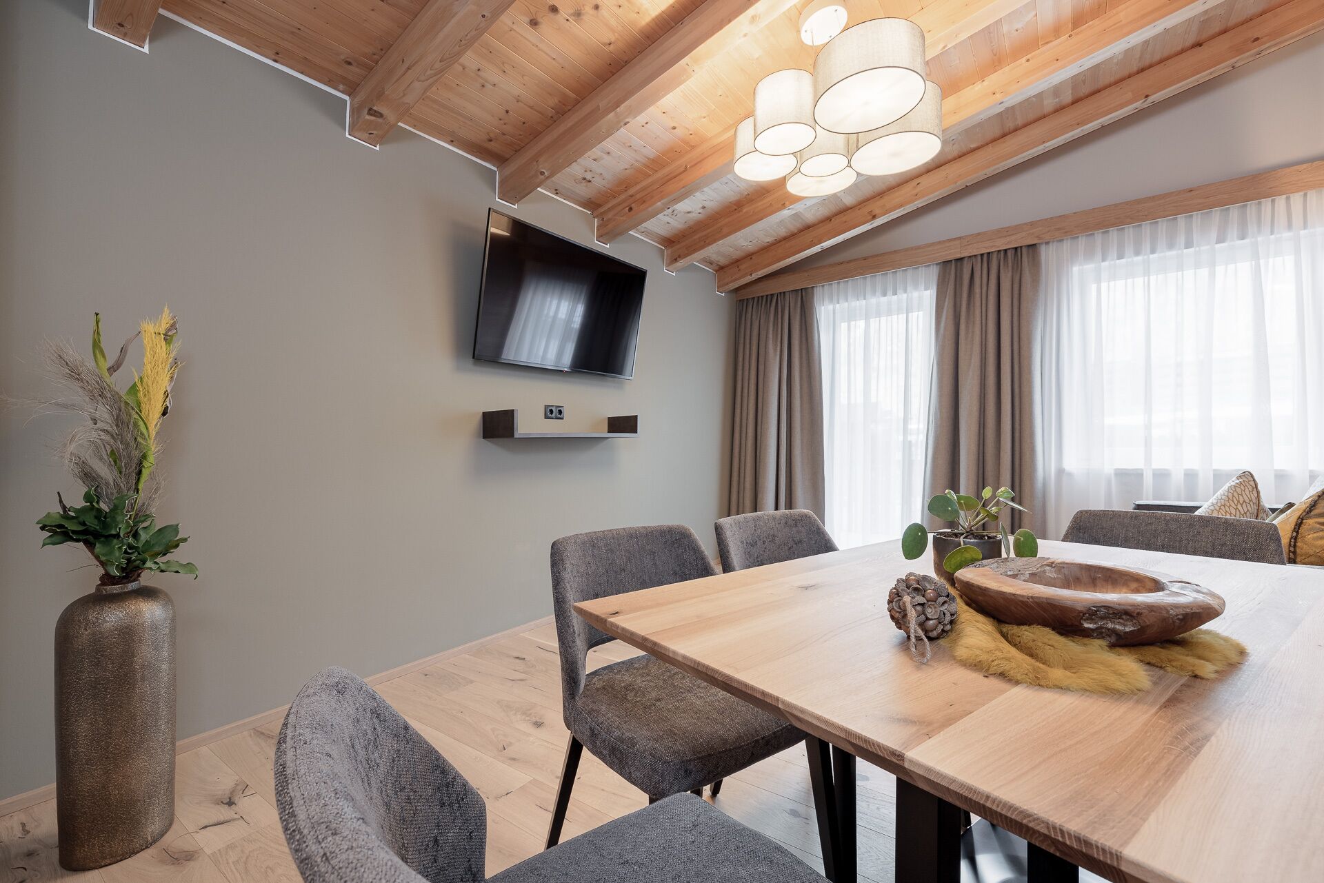 Un comedor moderno con una mesa de madera, sillas grises tapizadas, un televisor montado y un gran ventanal con cortinas.