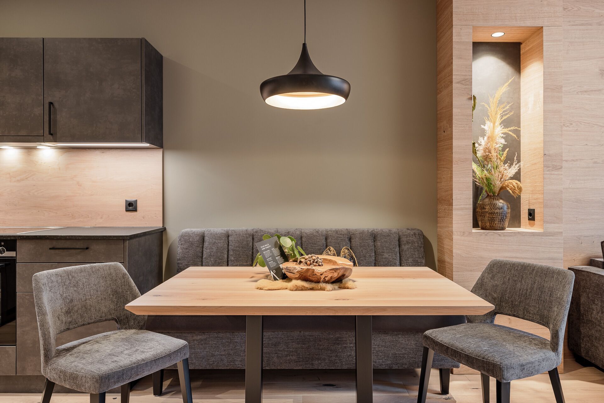 Un comedor moderno con una mesa de madera, sillas grises tapizadas, una lámpara colgante y un jarrón decorativo en la pared.