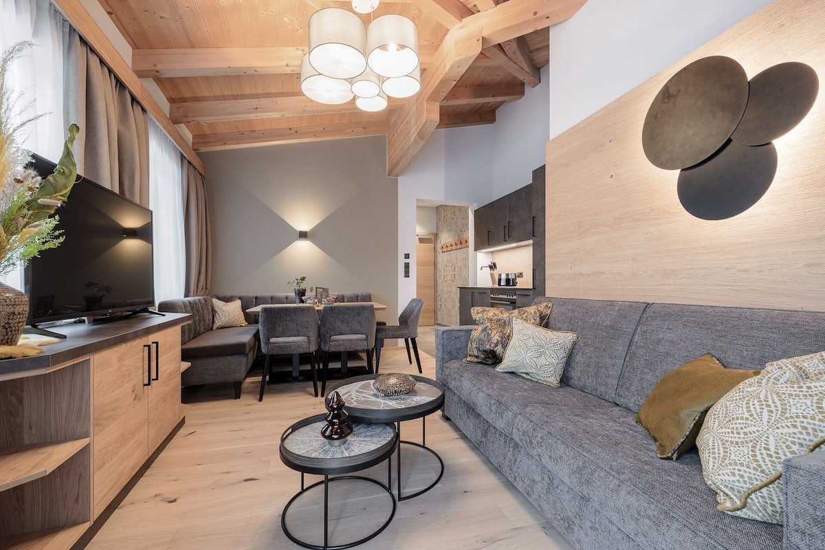 Ein modernes Wohnzimmer mit grauem Sofa, einem Esstisch und einer Küchenzeile mit Holzschränken und einem Einbauofen.