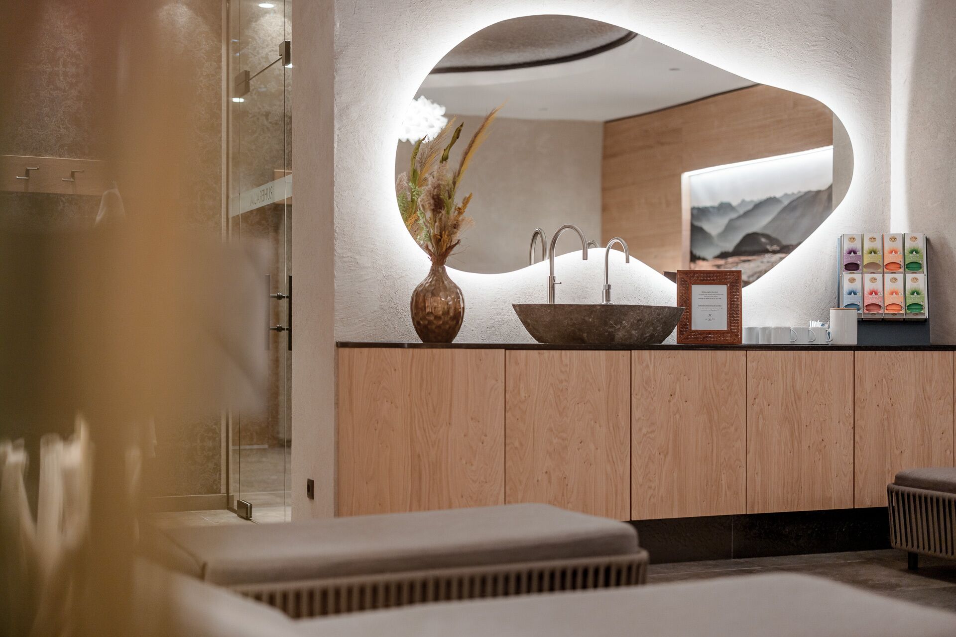 A modern bathroom with a large mirror, a wooden vanity, a sink with a bowl, and a vase with dried flowers.