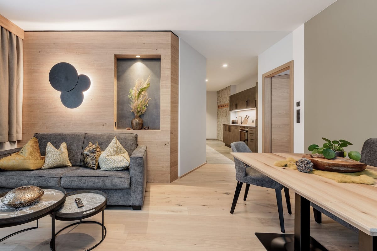 Ein modernes Wohnzimmer mit einem grauen Sofa, einem Holzessttisch und einer Küche im Hintergrund.
