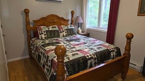 Una cama con dosel de madera con colcha, un equipo de aire acondicionado arriba, una ventana con cortinas y una mesita de noche con lámpara.