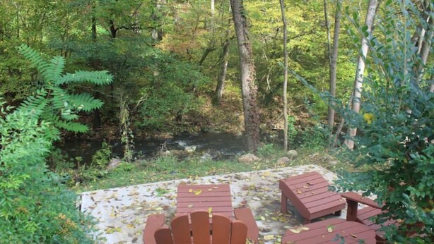 Una terraza de madera con sillas Adirondack y una mesa, rodeada de un bosque denso y un arroyo.