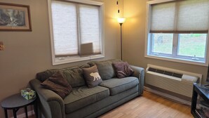 Una sala de estar con un sofá gris, dos ventanas con persianas, una lámpara de pie y un letrero colgado en la pared.