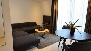 A modern living room with a sectional sofa, a small round coffee table, a TV stand, and a potted plant. - Apartment 22 (München)