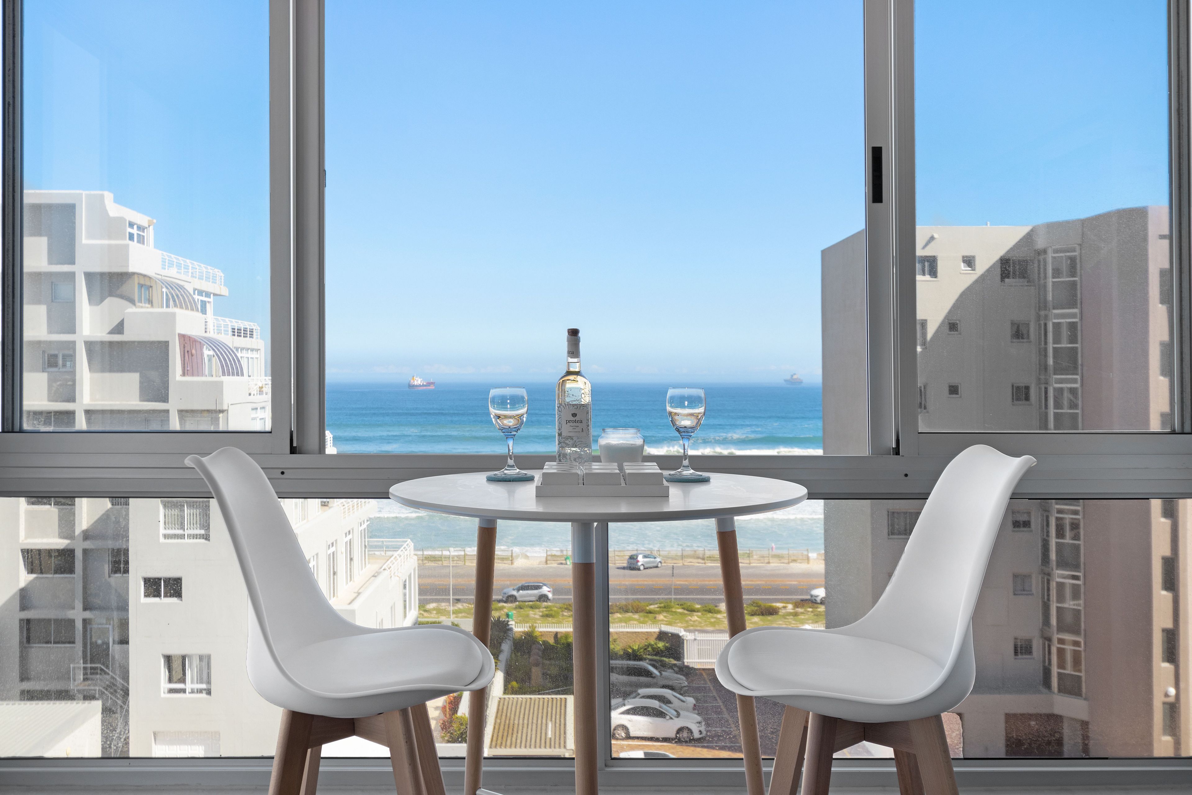 Habitació moderna amb una finestra gran que ofereix vista al mar i als edificis, amb dues cadires blanques, una taula petita amb una ampolla i gots.