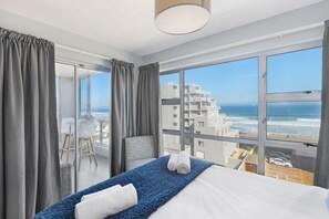 A hotel room with a large window offering a view of the ocean and buildings. There is a bed with a blue blanket and white pillows, a chair, and a small table with a lamp. - Ocean View 503 (Cape Town)