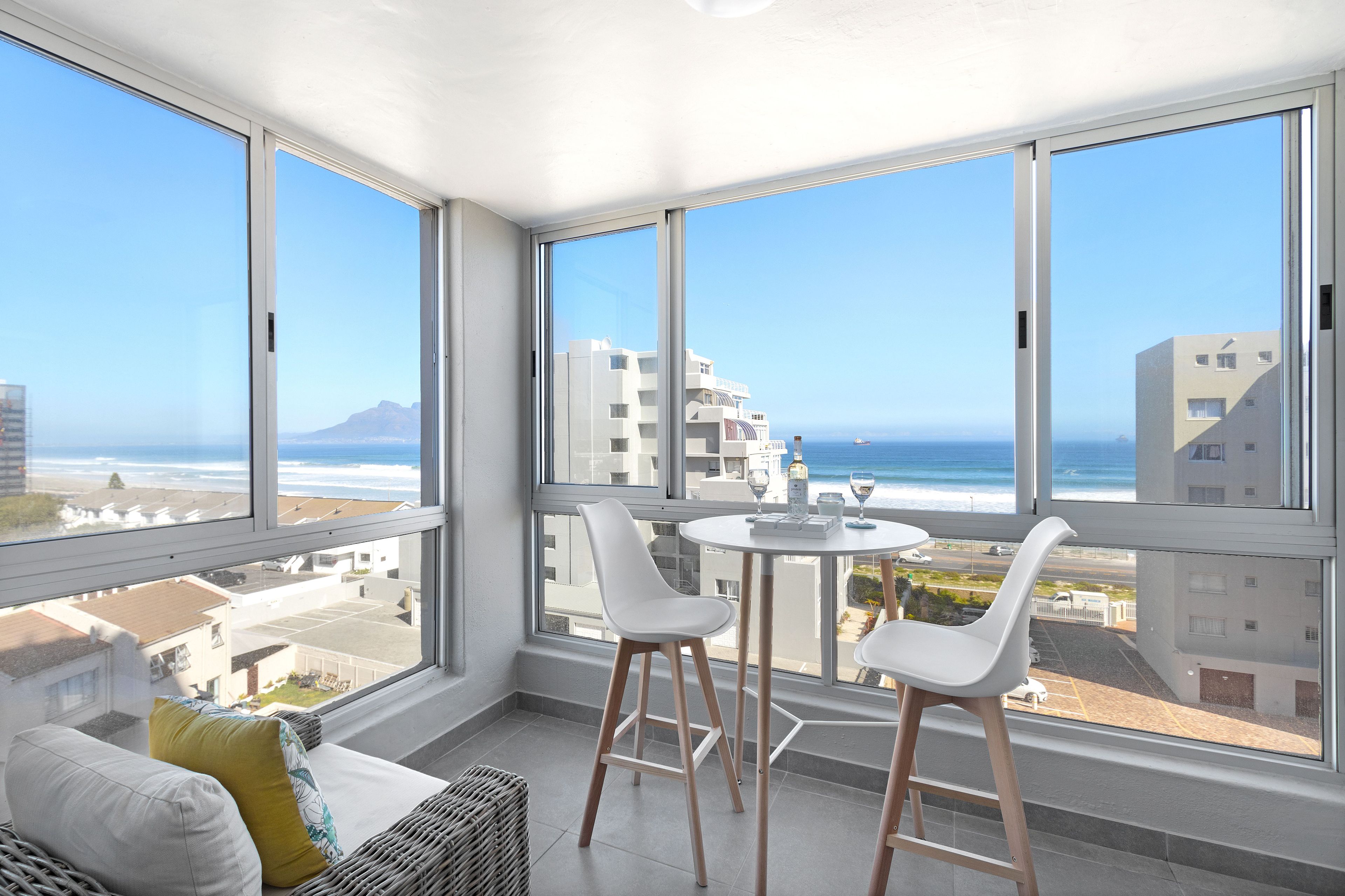 A balcony with a view of the ocean and buildings, featuring a white table and two white chairs.