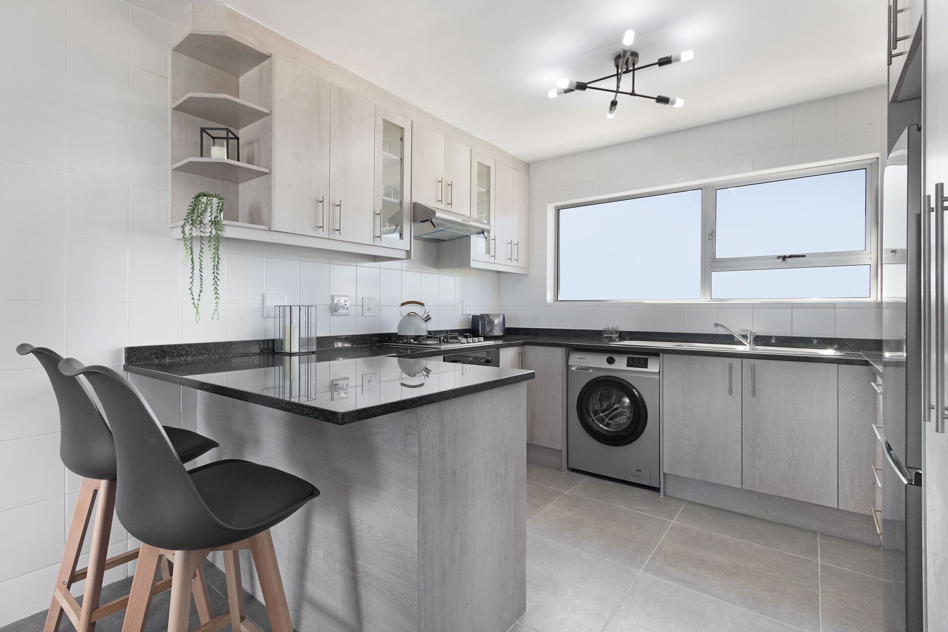 Una cocina moderna con una isla central, taburetes de bar, lavadora y un amplio ventanal que proporciona luz natural.