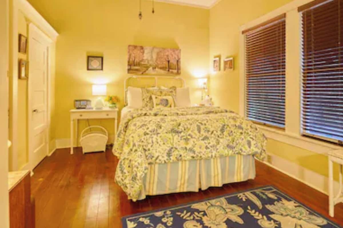 A bedroom with a bed, bedside tables, a nightstand, a window with blinds, and a door.