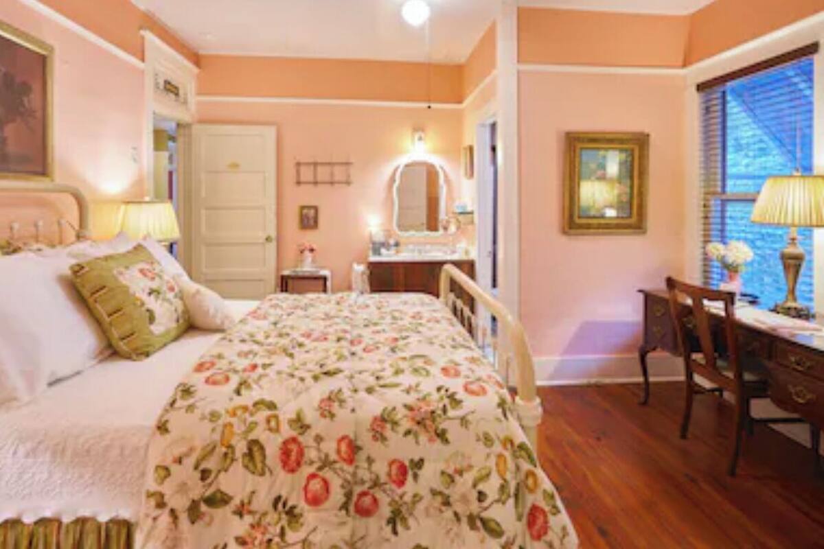 A bedroom with a floral bedspread, a bedside table, a mirror, a desk with a lamp, and a chair.