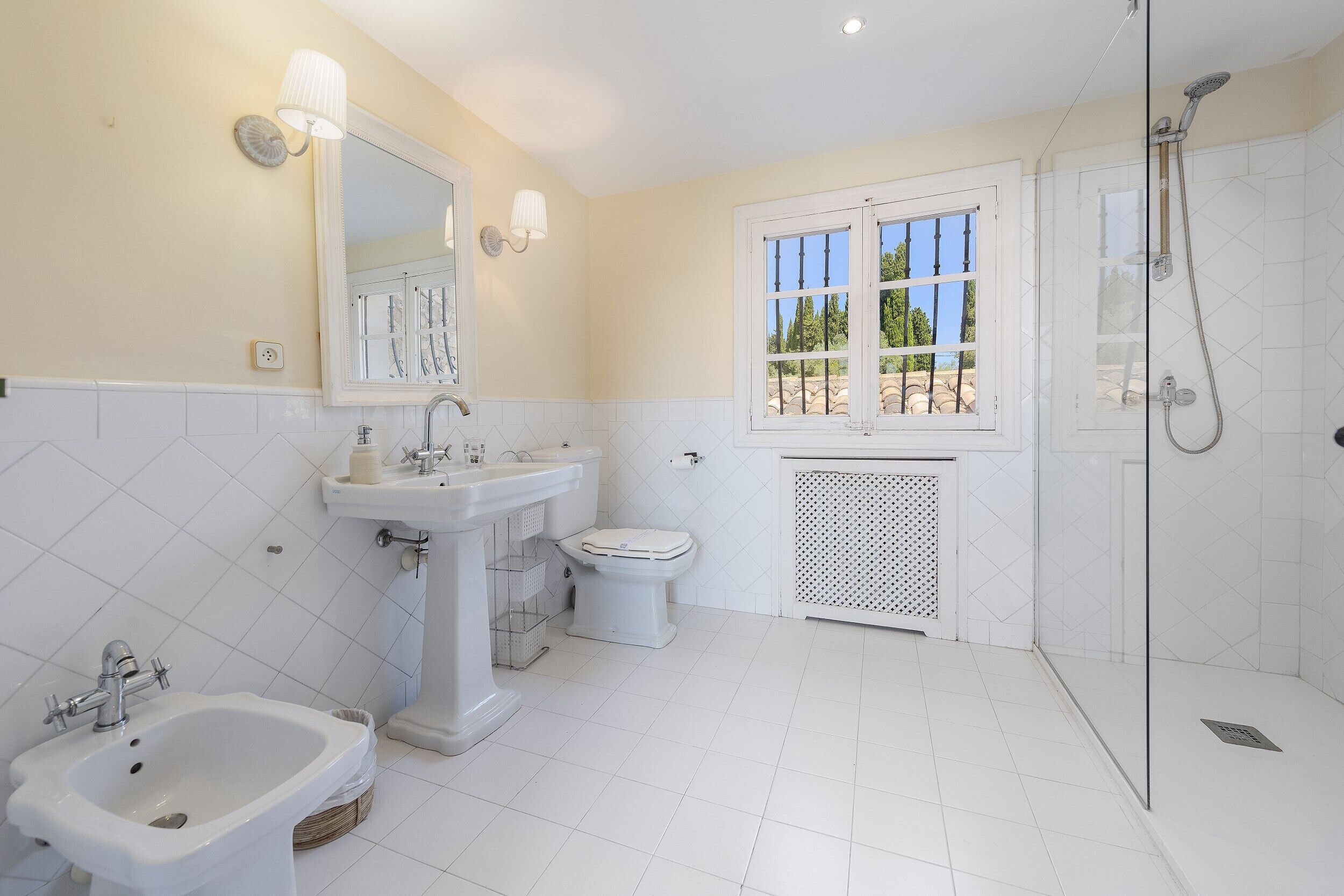 Un baño con lavamanos pedestal, inodoro, bidé, ducha y ventana con vista a árboles.