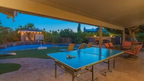An outdoor ping pong table with chairs, a pool, and a covered seating area with a view of a house and trees. - Entertainer's Dream Home! 3 Bdrm, Pool, Games! (Phoenix)