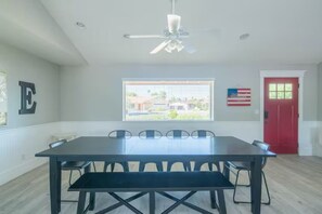 Un comedor con mesa y sillas, un ventilador de techo, una ventana con persianas y una puerta roja.