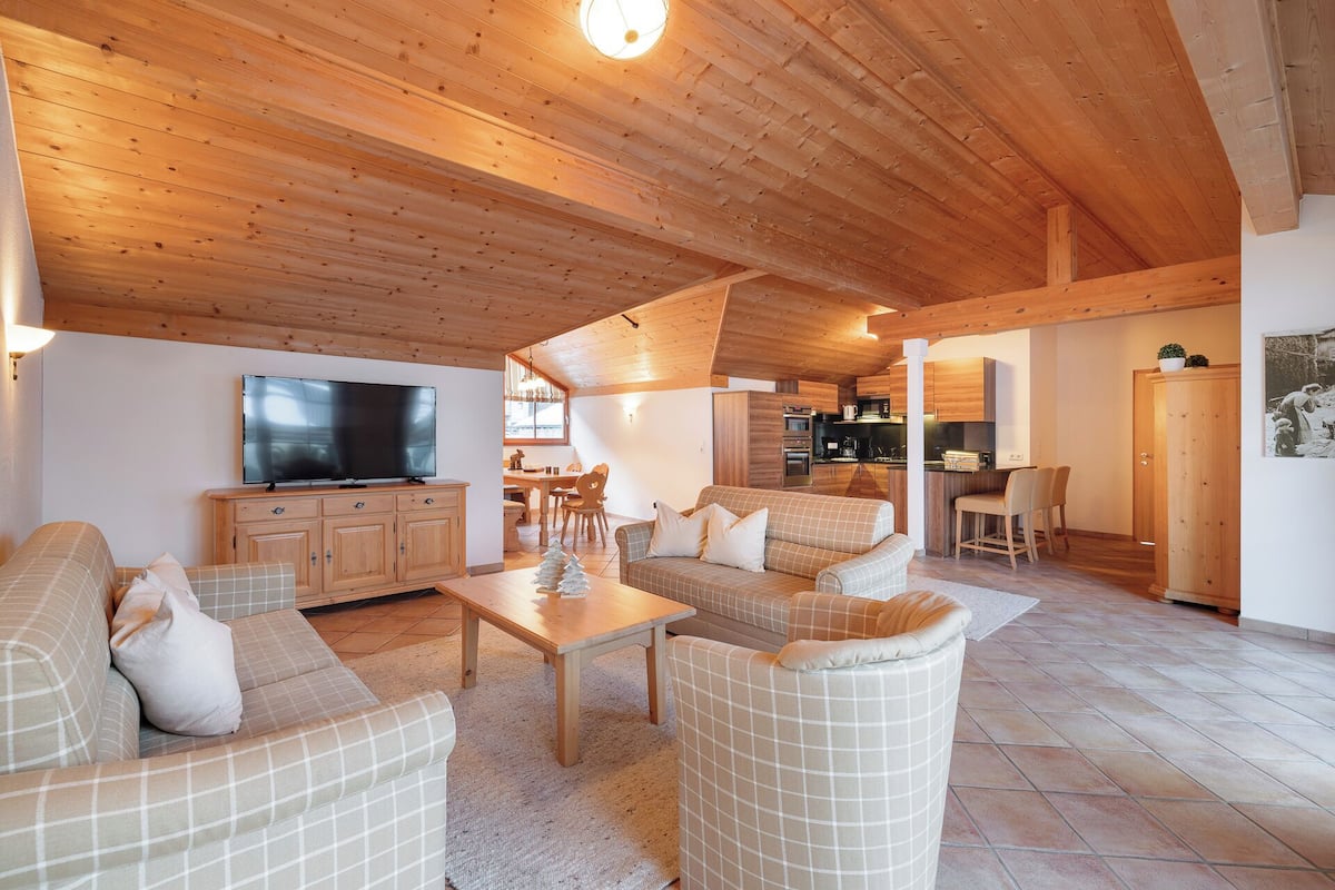 A spacious living area with a high ceiling, wooden furniture, a flat-screen TV, and a kitchen in the background.