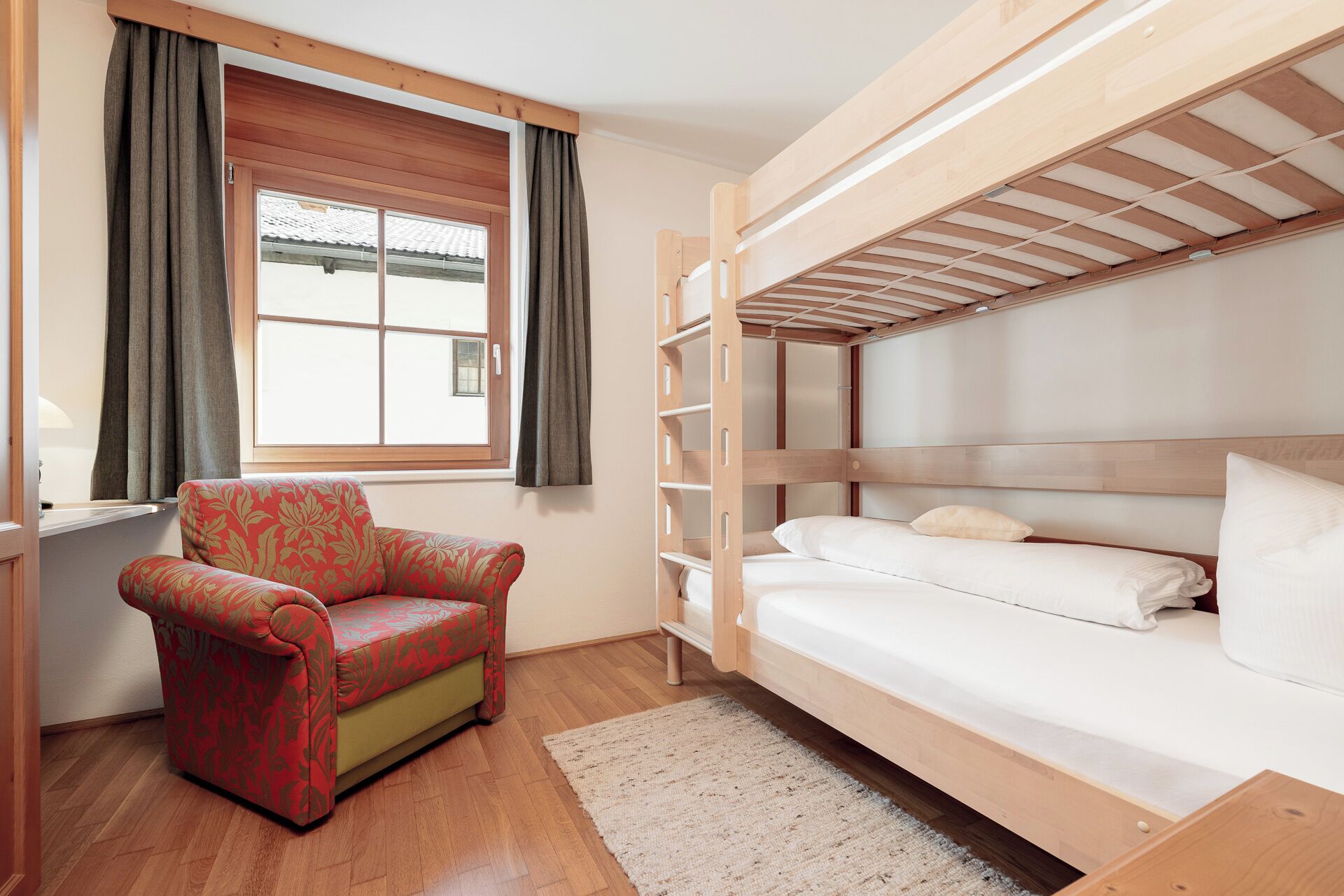Un letto a castello con struttura in legno e biancheria bianca, una poltrona rossa con motivo decorativo e una finestra con tende.
