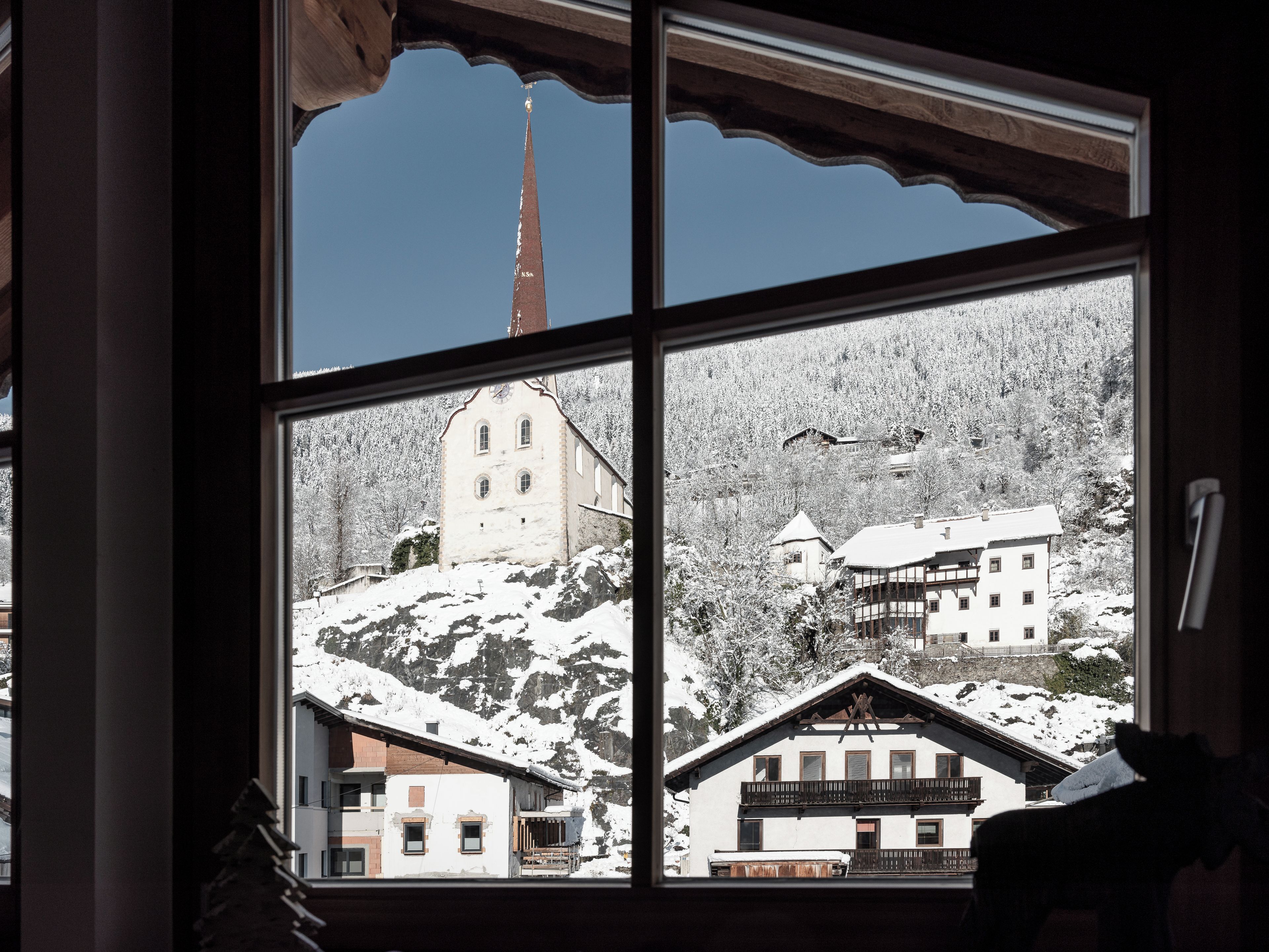 A window view of a snow-covered mountain village with traditional buildings and a church steeple.