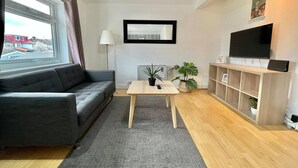 A modern living room with a grey sofa, a wooden coffee table, a TV on a wooden shelf, and a large window with curtains. - Lovely 2-bed Tree-hole in Northolt Southharrow (Northolt)