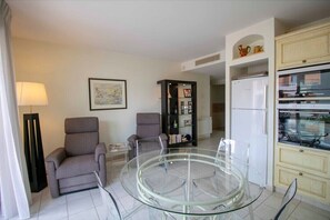 A modern living room with a glass coffee table, two armchairs, a bookshelf, and a kitchen area with a refrigerator.