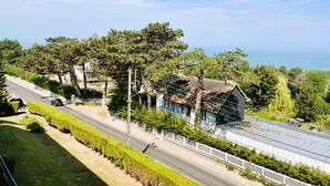 A coastal road with a modern building featuring a large glass facade and a metal roof, surrounded by lush greenery and trees. - Aux Flots Bleus,  avec balcon et vue sur mer (Trouville-sur-Mer)