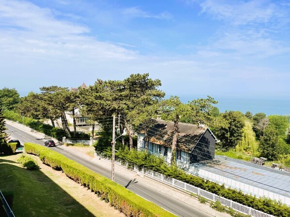 A coastal road with a modern building featuring a large glass facade and a metal roof, surrounded by lush greenery and trees. - Aux Flots Bleus, avec balcon et vue sur mer (Trouville-sur-Mer)