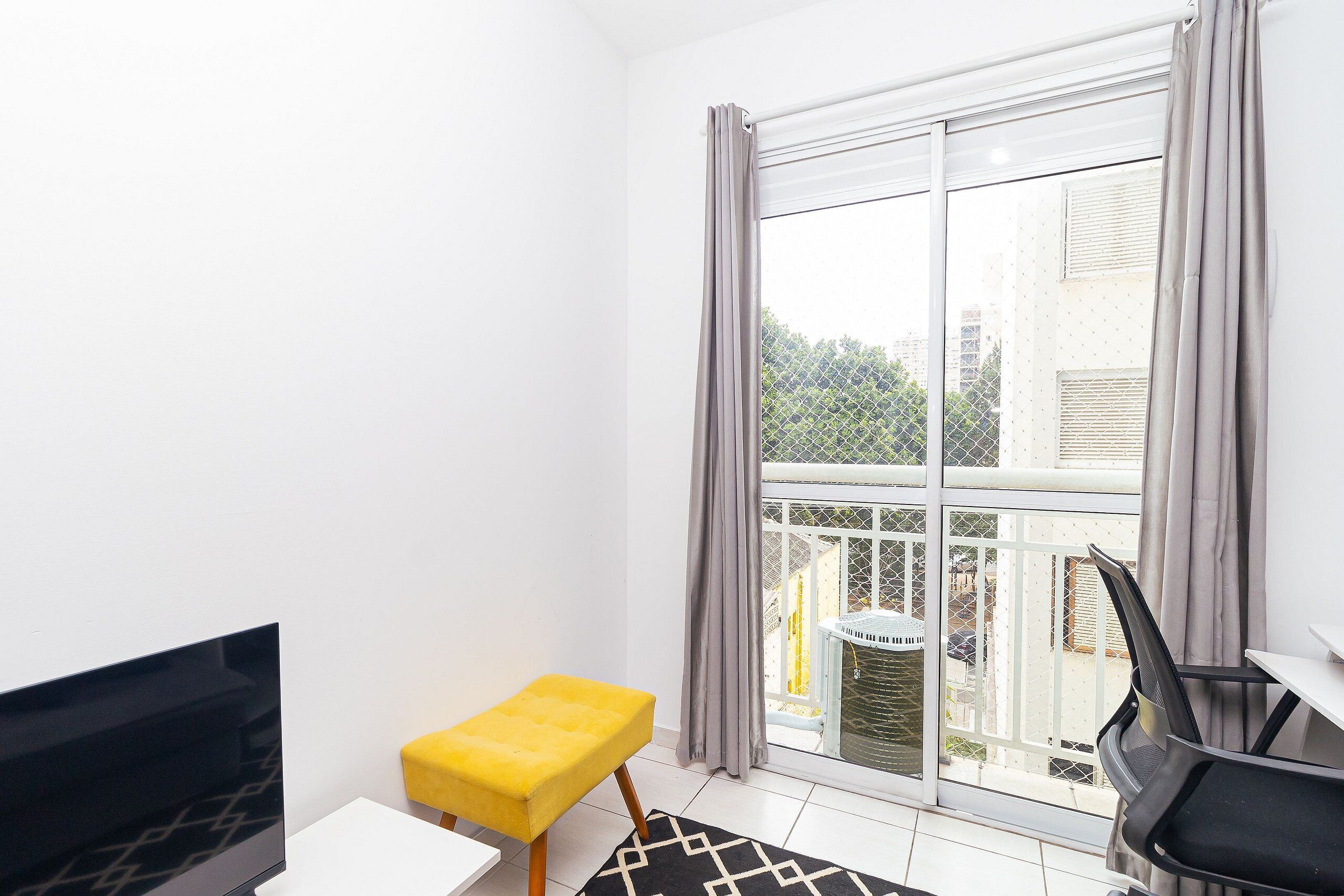 A small room with a TV, a yellow ottoman, a black and white rug, and a balcony with a view of buildings and greenery.